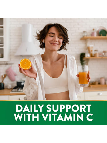 Young adult woman smiling, holding an orange and orange juice in a kitchen. Text 'DAILY SUPPORT WITH VITAMIN C' is at the bottom.