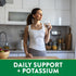 Young adult woman drinks water in a modern kitchen. Green banner reads 'DAILY SUPPORT + POTASSIUM'.