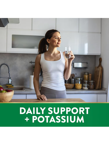 Young adult woman drinks water in a modern kitchen. Green banner reads 'DAILY SUPPORT + POTASSIUM'.