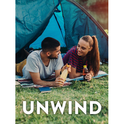 Young adult man and woman camping, relaxing on grass outside a blue tent, holding drinks. Text: UNWIND.