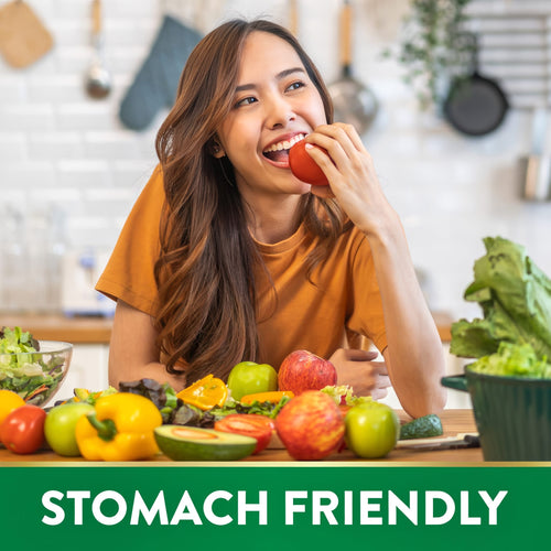 Young woman happily eating a tomato, surrounded by fresh fruits and vegetables on a kitchen counter. Text banner reads 'STOMACH FRIENDLY'.