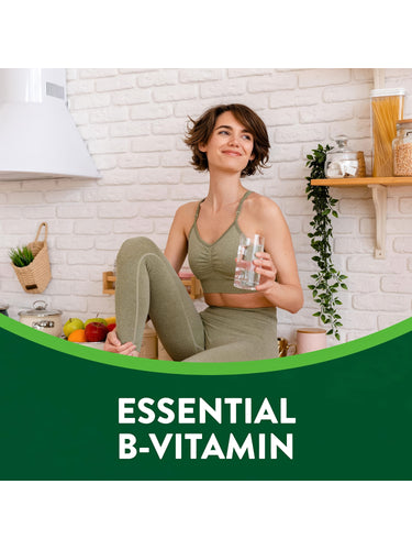 Young adult woman in activewear sits in a kitchen holding a glass of water with 'ESSENTIAL B-VITAMIN' text in view.