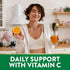 Young adult woman smiling, holding an orange and orange juice in a kitchen. Text 'DAILY SUPPORT WITH VITAMIN C' is at the bottom.