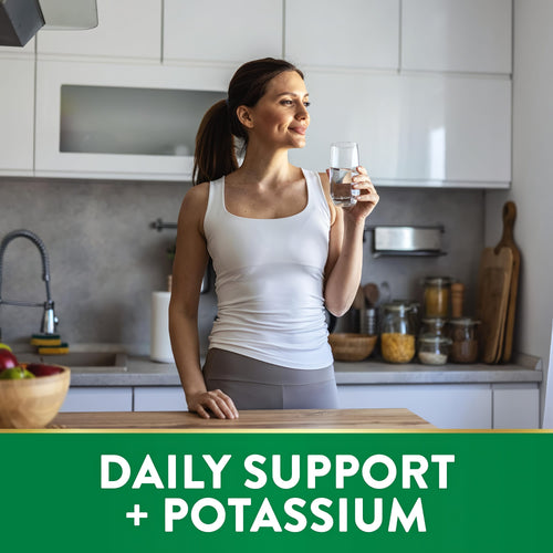 Young adult woman drinks water in a modern kitchen. Green banner reads 'DAILY SUPPORT + POTASSIUM'.