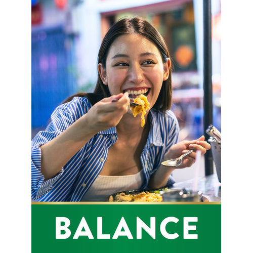 Young Asian woman laughing while eating pasta, with a green graphic displaying 'BALANCE', at a restaurant table.