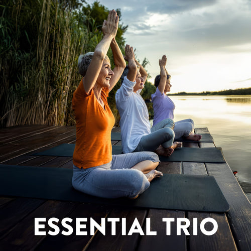 Three senior women practicing yoga with raised arms on mats by a lake at sunset. Text: ESSENTIAL TRIO.