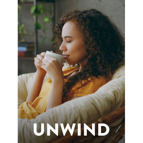 A young woman with curly hair, eyes closed, sits in a chair, sipping from a white mug with the text 'UNWIND'.