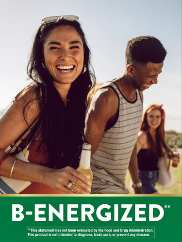 A young woman smiles, holding a 'B-ENERGIZED' bottle, while friends socialize outdoors.